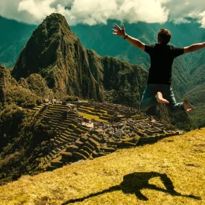 A man jumping for joy on a grassy hill overlooking the ancient stone ruins of Machu Picchu with Huayna Picchu mountain in the background.