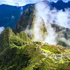 Panoramic view of the stone structures of Machu Picchu with thick white clouds swirling around the surrounding green mountains and Huayna Picchu peak.